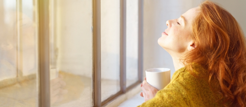 Blissful Young Woman Enjoying The Sun On Her Face As She Leans On A Window Sill With Her Head Tilted Back And A Mug Of Coffee