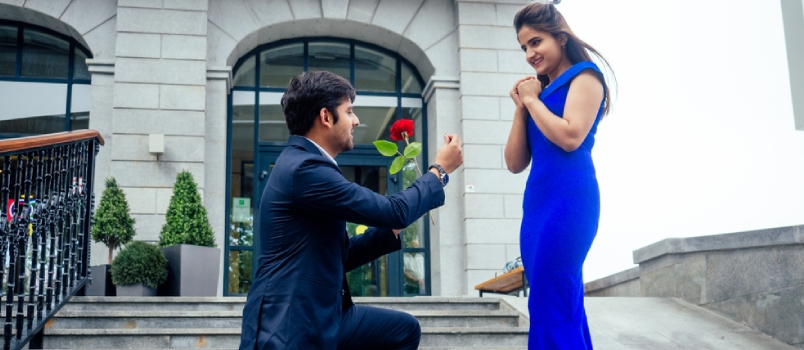 Happy Asian Man In Stylish Black Suit Falling Knee In Front Of His Beautiful Woman In Blue Long Dress Asking Proposal And Giving Gold Ring On The Background Of The Restaurant Street
