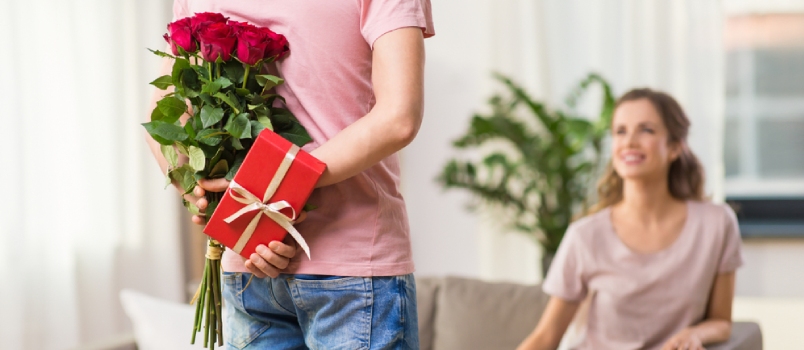 Happy Woman Looking At Man Hiding Flowers And Gift Behind His Back At Home