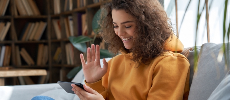 Happy Young Hispanic Latin Teen Girl Sit On Sofa At Home Holding Phone Looking At Screen Waving Hand On Video Calling