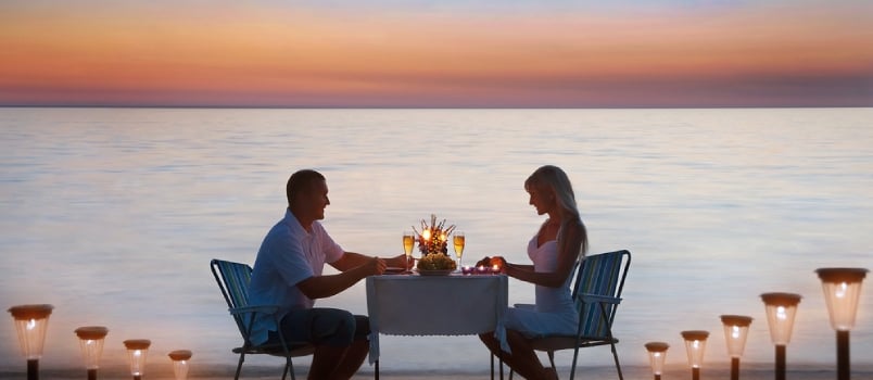 Loving Couple Share A Romantic Dinner With Candles Lanterns And Wine Glasses At Sea Beach Sand Against Wonderful Sunset