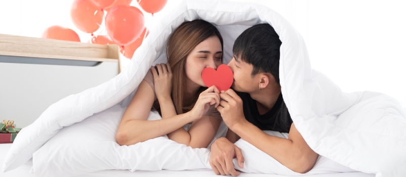 Happy Young Couples Hold And Kiss A Red Heart Together On A Bed Under A Blanket On Morning In The Bedroom