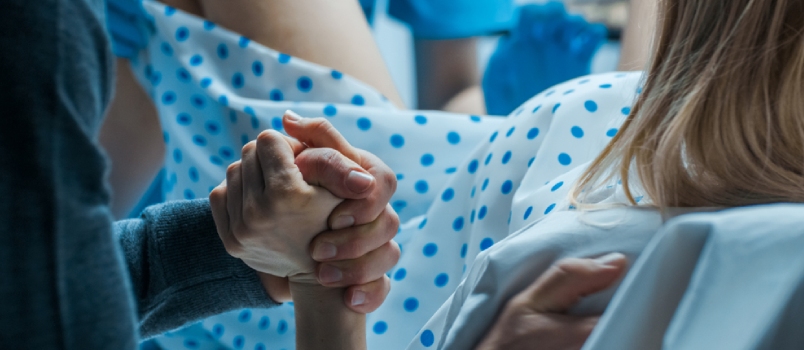 Emergency In The Hospital: Woman Giving Birth, Husband Holds Her Hand In Support, Obstetricians Assisting. Modern Delivery Ward With Professional Midwives.