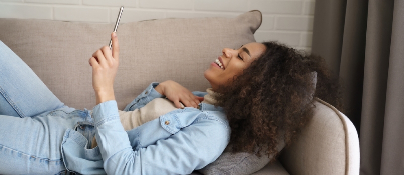 Happy Afro American Young Woman Laughing Watching Funny Video On Smartphone Lying On Sofa At Home