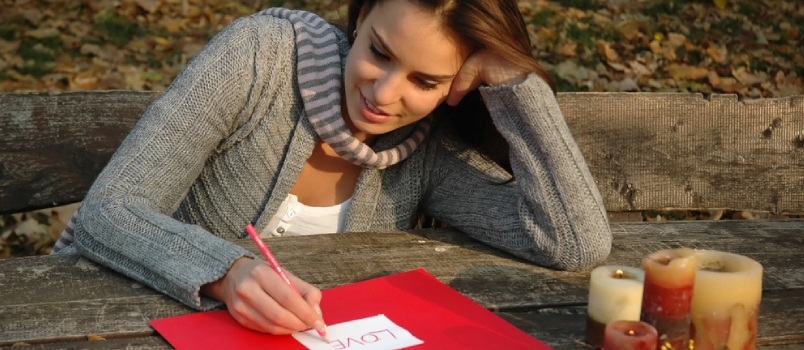Woman Thoughtfully Smiling, Leaning On The Arm, Writing Word Love On White And Red Paper On Wooden Table With Candles