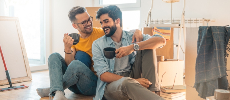 Cute Gay Couple Sitting On Floor And Enjoying Coffee In New Home