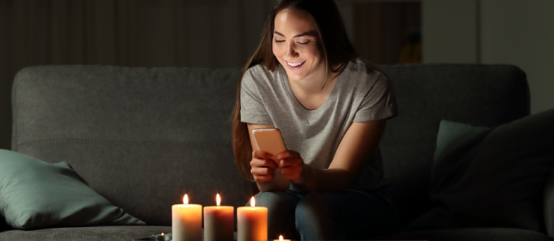 Woman using a smart phone in the night with candle lights sitting on a couch in the living room at home