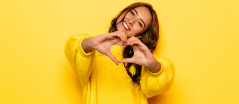 Smiling Young Girl In Yellow Sweater Showing Heart With Two Hands, Love Sign. Isolated Over Yellow Background.