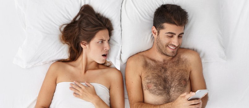 Men And Women Lying Together On Bed, Men Using Cellphone While Irritated Women Staring At Husband 