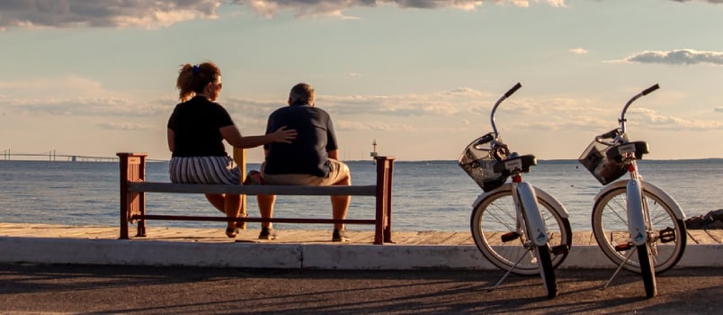 A Middle Aged Caucasian Couple Is Sitting On A Bench By The Beach. They Have Their Identical Bikes Parked Next To Them
