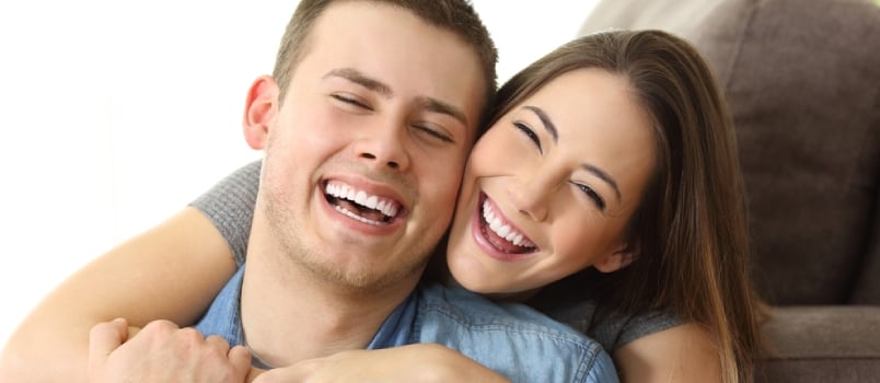 Happy Couple With Perfect White Smile Posing And Looking At Camera On A Couch At Home