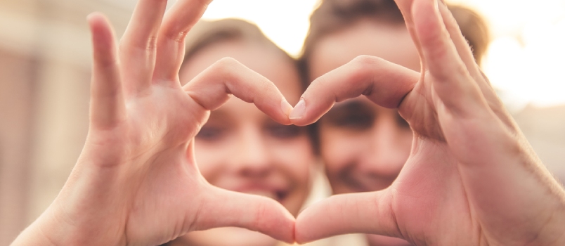 Beautiful Young Couple Is Making Heart Of Fingers, Looking Through It And Smiling, Hands In Focus, Close-up