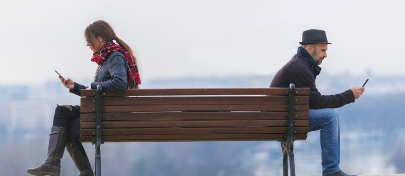 Man And Woman Sitting Apart On A Park Bench And Using Mobile