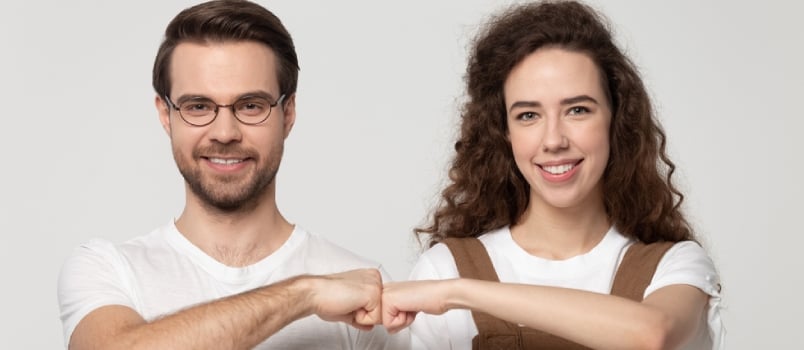 Happy Young Man In Eyewear Bumping Fists With Millennial Smiling Girl, Isolated On Grey White Studio Background