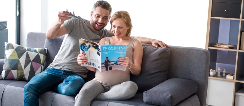 Planning Future Journey. Full Length Portrait Of Cheerful Loving Couple Reading Magazine About Traveling While Sitting On Sofa At Home