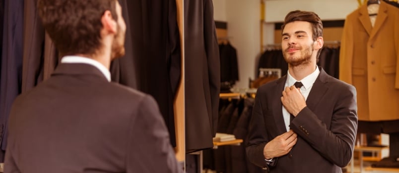 Modern Young Handsome Businessman Dressed In Classical Suit Adjusting A Tie In Front Of The Mirror While Standing In The Suit Shop