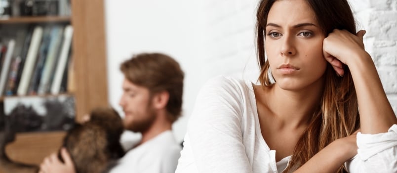 Upset Thoughtful Women Worried About Something While Men Playing With The Toy Of Pet Behind Sitting On Sofa At Living Room