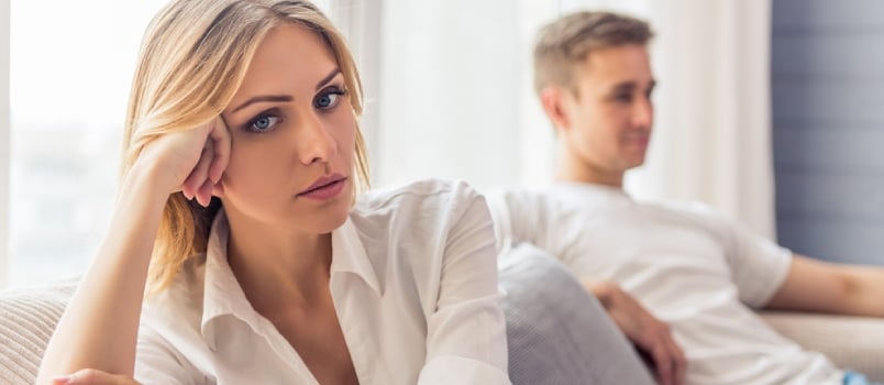 Beautiful Young Woman Is Sitting Sad On Couch At Home, Her Boyfriend Is Sitting In The Background