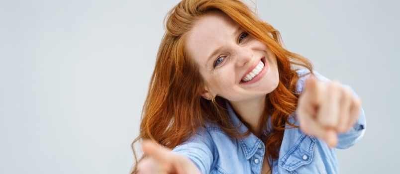 Smiling Happy Woman Pointing At The Camera With Both Hands And Her Head Tilted To The Side With A Playful Expression And Focus To Her Face Over Grey