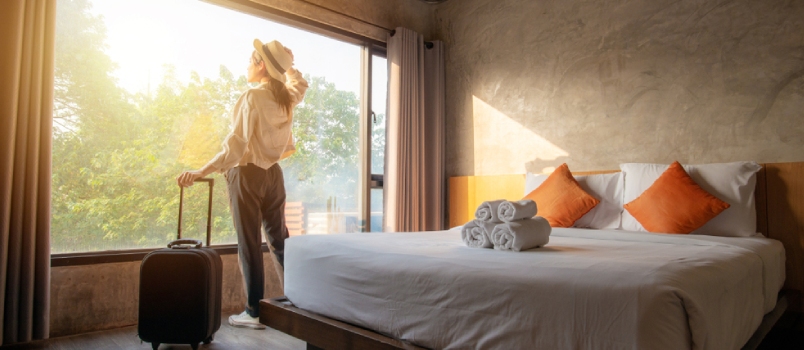 Portrait Of Tourist Woman Standing Nearly Window, Looking To Beautiful View With Her Luggage In Hotel Bedroom