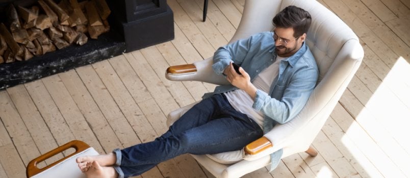 Relaxed Casual Guy Lounge Sit on Comfortable Armchair in Modern House Room With Wooden Floor Fireplace Using Smartphone Apps