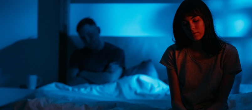 Scared Insecure Humiliated Woman Sitting on Bed on Distance From Aggressive Man in Dark Night Red Blue Neon Light on Defocused Background