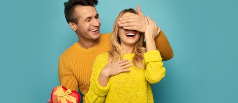 A Nice Man With Raven Black Hair Is Covering the Eyes of His Girlfriend, Looking at Her With a Big Smile and Holding a Red Heart-shaped Box in His Right Hand