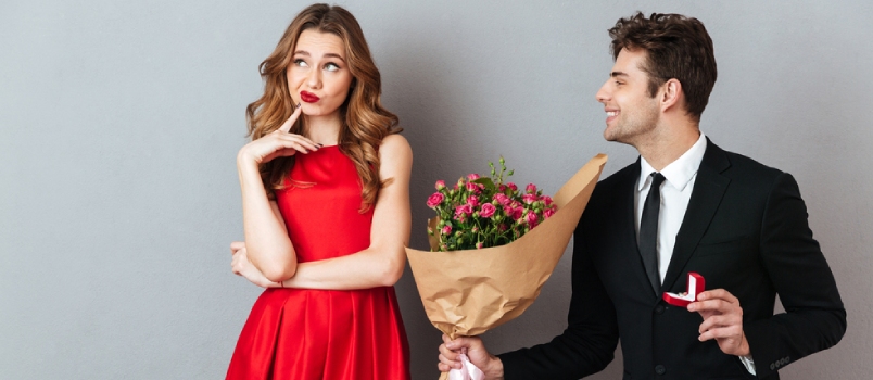 Portrait of a Joyful Man Proposing to a Unsatisfied Girl With Flowers and an Engagement Ring Over Gray Wall Background