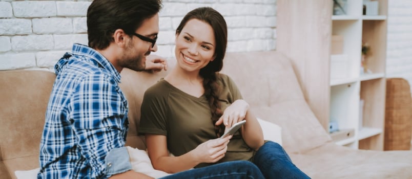 Girl Shows Man Smartphone. Happy Family. Male and Famale in Brown Sofa. White Pillows in Brown Sofa.