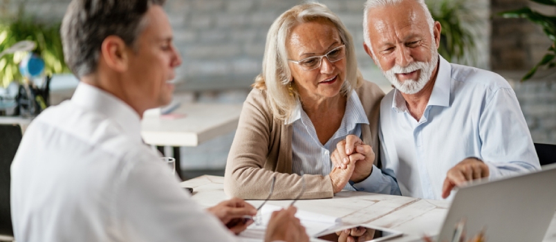 Happy Senior Couple Holding Hands and Using Laptop While Having a Meeting With Financial Advisor in the Office