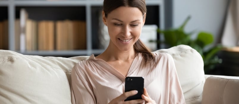 Smiling Young Woman Using Phone, Sitting on Couch at Home, Looking at Smartphone Screen