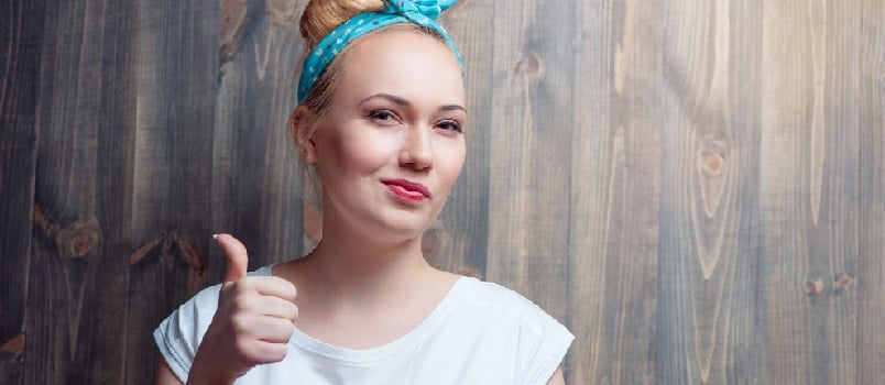 Young blond woman showing thumb up with sceptic grimace against wooden wall.