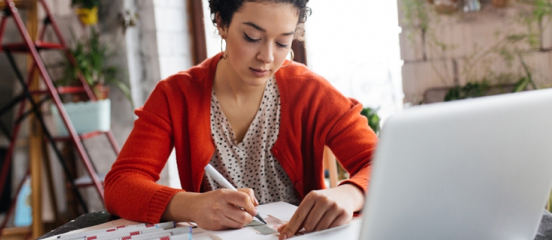 Young Beautiful Woman With Dark Curly Hair Sitting at the Table With Laptop Dreamily Drawing Fashion