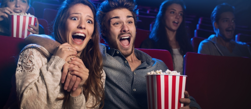 Young scared couple at the cinema watching an horror movie and screaming, she is holding her boyfriend's hand