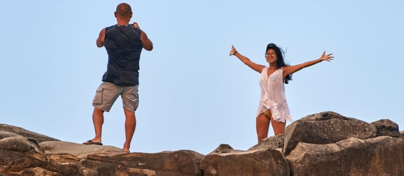 A woman poses with arms outstretched joyfully, as her partner takes her photograph