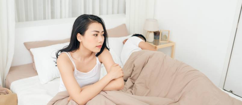 Unhappy woman sitting on bed while man is sleeping