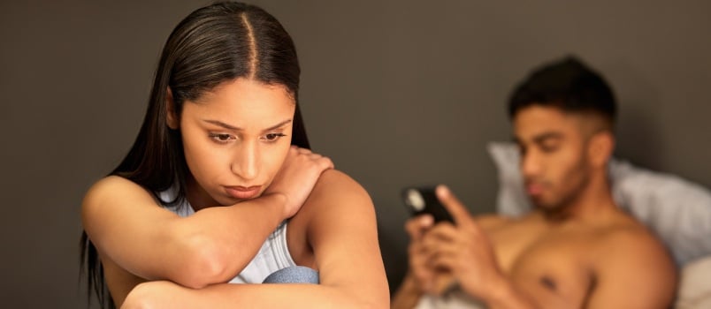 Upset girlfriend sitting on bed while boyfriend is using cellphone