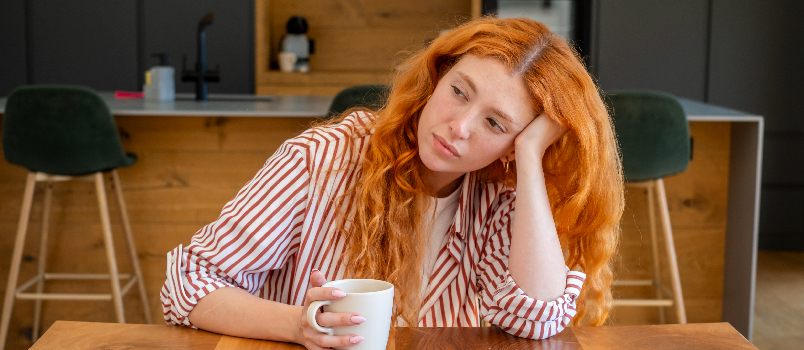 Pensive Young Woman With Red Hair Sitting in Modern Kitchen
